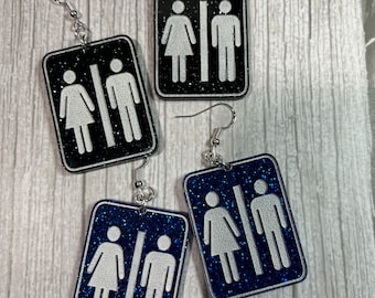 May include: A pair of earrings with a black and blue glitter background. The earrings feature a white silhouette of a male and female restroom sign.
