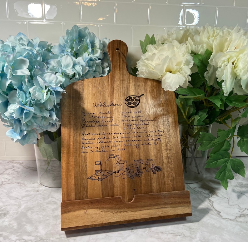 May include: A wooden cutting board with a recipe for Uebleskever, a type of Dutch pancake, engraved on the surface. The recipe includes ingredients like milk, margarine, salt, sugar, and yeast. The board has a built-in stand for propping it up.