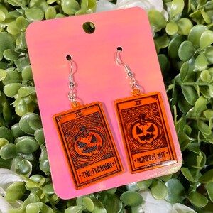 May include: Orange acrylic earrings featuring a jack-o'-lantern design within a tarot card frame. The text "THE PUMPKIN" and "XIX" are visible. The earrings hang from silver-colored hooks, perfect for Halloween.