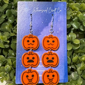 May include: A pair of orange pumpkin dangle earrings with different facial expressions. The earrings are on a blue and white card with the text "Whimsical Craft Co."