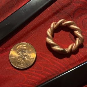 May include: A copper-colored ring with a twisted rope design sits next to a U.S. penny. The ring is made of a metal that appears to be copper or a copper alloy. The penny is a standard one-cent coin, showing Abraham Lincoln.