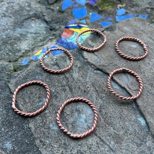 May include: Six copper-colored rings with a twisted rope design are arranged on a gray stone surface. The rings are circular and appear to be handmade. The rings are a warm, reddish-brown color.