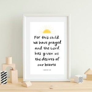 May include: A framed print with the text "For this child we have prayed and the Lord has given us the desires of our hearts." The print includes a yellow sun illustration and the Bible verse 1 Samuel 1:27. The frame is white.