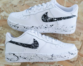 May include: White leather trainers with a black swoosh logo and black paint splatters on the sole and swoosh. The trainers have a white midsole and a black outsole.