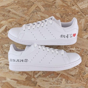Peut inclure: Une paire de baskets en cuir blanc avec un cœur rouge et le texte "mtG" sur une chaussure et le texte "22.06.2024" et deux anneaux sur l'autre chaussure.