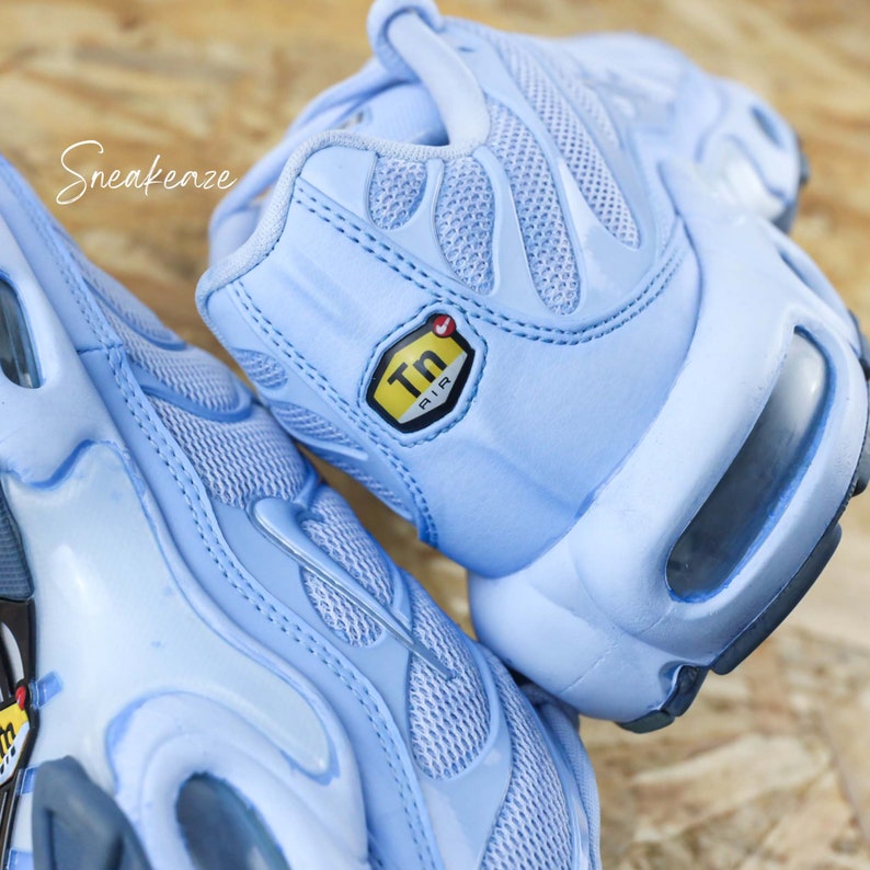 May include: A pair of light blue Nike Air Max Plus sneakers with a yellow "Tn Air" logo on the side. The sneakers have a mesh and synthetic upper and a visible air unit in the heel.