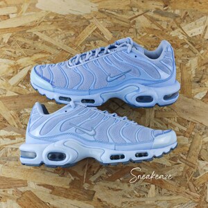 May include: A pair of light blue Nike Air Max Plus sneakers with a mesh upper and a black and white sole. The sneakers are on a wooden surface.