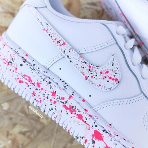 May include: White sneakers with a pink, black, and tan splatter paint design. The Nike swoosh and midsole feature the same paint splatter pattern. The word "sneakeaze" is printed on the side of the shoe.