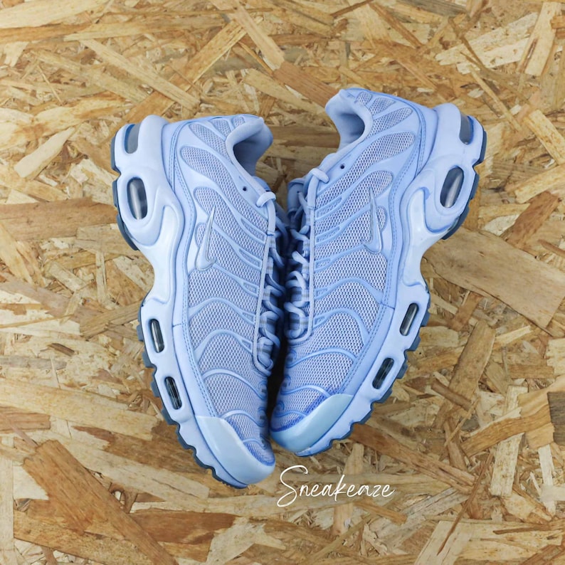 May include: A pair of light blue Nike Air Max Plus sneakers with a mesh upper and a white swoosh logo. The shoes are on a light brown wooden surface.