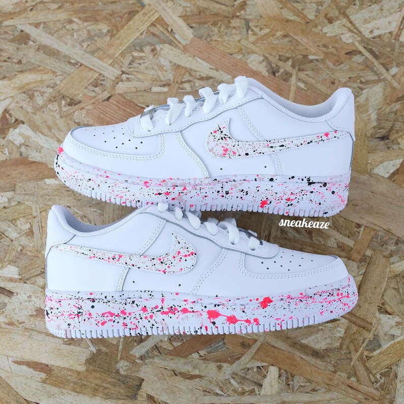 May include: Two white sneakers with pink and black paint splatter details on the soles and Nike swoosh. The shoes have white laces and the word "sneakeaze" is printed on the side.