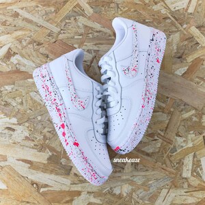 May include: White sneakers with pink and black paint splatter design. The shoes have white laces and a Nike swoosh. The word "sneakeaze" is visible on the side of the shoe.