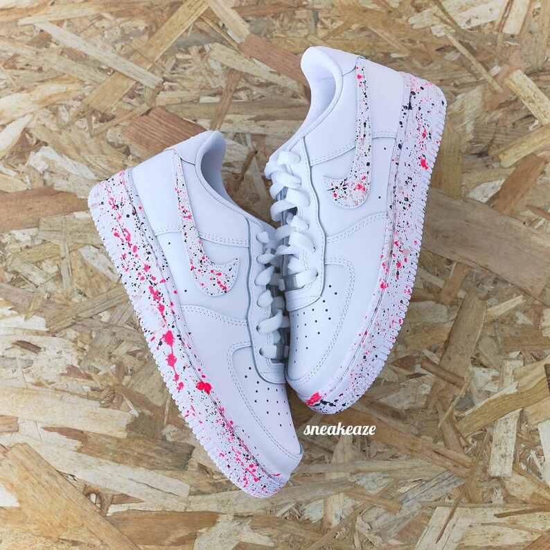 May include: White sneakers with pink and black paint splatter design. The shoes have white laces and a Nike swoosh. The word "sneakeaze" is visible on the side of the shoe.