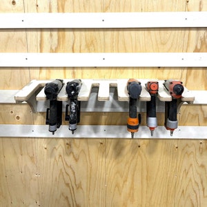 May include: A white wooden wall-mounted rack with six slots for holding tools. The rack is filled with six different power tools, including a black and silver nail gun, a black and orange nail gun, and a black and red nail gun.