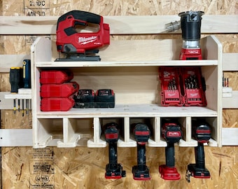 May include: A white wooden wall-mounted storage rack with red and black power tools. The rack has two shelves and a bottom row of hooks. The tools include a red and black jigsaw, a red and black drill, and a red and black battery pack. The rack is mounted on a wood panel wall.