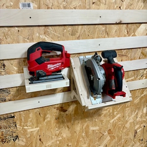 May include: Two power tools, a red and black Milwaukee FUEL jigsaw and a circular saw, mounted on wooden shelves against a wood-paneled wall. The jigsaw is on the left, and the circular saw is on the right.
