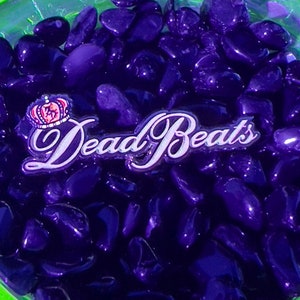 May include: A neon green glass dish filled with dark purple stones. The dish has a decorative edge and contains the text "Dead Beats" in a cursive font. A small crown emblem is also visible.