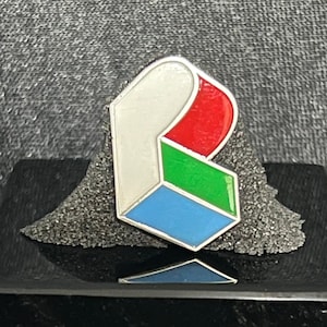 May include: A silver pin with a stylized letter B design. The letter is made up of four colored blocks: white, red, green, and blue.
