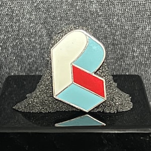 May include: A white, blue, and red enamel pin shaped like the letter "B". The pin is a geometric design with a three-dimensional effect.