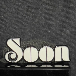 May include: White enamel pin with the word "Soon" in a retro font. The pin is on a black surface.