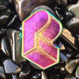 May include: A glittery, iridescent letter "R" pin with a geometric design. The pin features a gradient of pink and gold hues, outlined with a rainbow-colored border. The pin is set against a background of polished black stones.