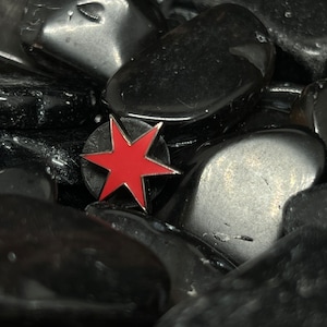 May include: A red star pin on a black background. The star is surrounded by black stones.