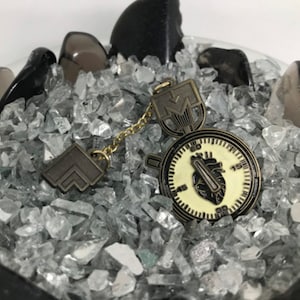 May include: A bronze-colored pendant with a clock face design and a heart graphic. The pendant is connected by a gold chain to a matching geometric charm. The items are displayed on a bed of clear, crushed glass.
