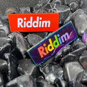 May include: Two rectangular pins with the word "Riddim" on them. The top pin is red with white lettering. The bottom pin is iridescent with rainbow lettering.
