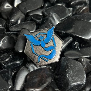 May include: A silver and blue enamel pin featuring a stylized bird with wings spread, set against a hexagonal background. The pin is surrounded by black stones.