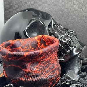 Puede incluir: Un cráneo negro con un tubo de tela con un patrón de flujo de lava rojo y naranja. El tubo de tela está hecho de un material suave y elástico y tiene una superficie texturizada.