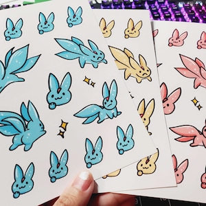 May include: Three sticker sheets featuring cartoon creatures with long ears and multiple tails in blue, yellow, and pink. The designs include various poses of these creatures, some with small star accents, printed on white paper with a glossy finish.