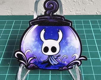 May include: A sticker featuring a cartoon character inside a purple and blue cauldron. The character has a white head with two horns and large black eyes. The cauldron is surrounded by black and purple foliage.