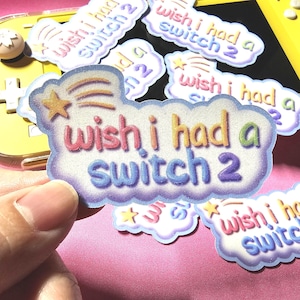 Könnte beinhalten: Ein glitzernder Aufkleber in Wolkenform mit einem Sternschnuppen-Symbol. Der Text "wish i had a switch 2" ist in einer mehrfarbigen Verlaufsschrift gedruckt. Der Aufkleber hat einen violetten Rand und eine texturierte, funkelnde Oberfläche.