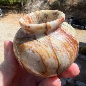 May include: A small, round decorative vase with a wide opening. The vase is made of a stone material with a swirling pattern of tan, brown, and cream colors. The vase is held in a hand.
