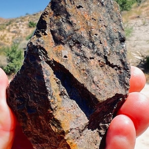 Peut inclure: Une roche brute de forme irrégulière, présentant un mélange de teintes marron foncé, orange et noires. La roche a une surface texturée avec de petits trous et une fissure diagonale prononcée. L'objet est tenu dans une main.