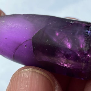May include: A large, purple, faceted gemstone with a rough, irregular shape. The gemstone is translucent and has a slight shimmer.