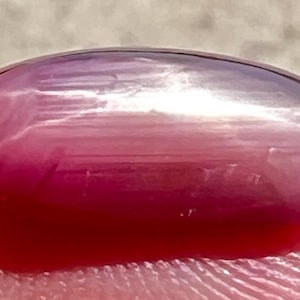 May include: A smooth, oval-shaped gemstone with a pink to white gradient. The gemstone has a pearlescent sheen and is a light pink color.