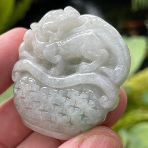 Könnte beinhalten: Ein weißer Jade-Anhänger, der mit einem detaillierten Design eines Drachen in einem Korb verziert ist. Der Anhänger ist glatt und poliert und hat einen dezenten Grünton.