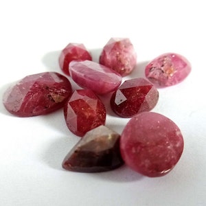May include: A collection of eight faceted ruby gemstones in various shapes and sizes. The gemstones are a deep red color and have a polished finish.