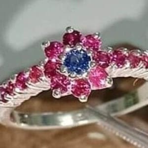 May include: A silver ring with a flower-shaped design featuring a blue sapphire surrounded by pink rubies.