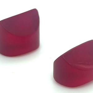 May include: Two translucent, ruby-colored cabochons with a rounded top and a flat bottom. The gemstones are cut into a half-cylinder shape, and are displayed against a white background.