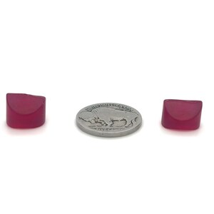 May include: Two small, translucent, ruby-colored, square-cut gemstones flank a silver coin. The coin features a buffalo on one side and text. The gemstones have a rounded top edge and a flat bottom.