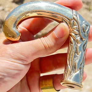 May include: A gold-toned metal walking stick handle with an ornate design. The handle is shaped like a curved hook and has a detailed floral pattern.