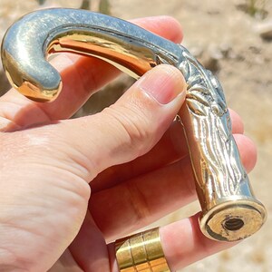 May include: A gold-toned metal walking stick handle with a detailed, carved design resembling a tree branch. The handle has a smooth, curved top and a threaded bottom.