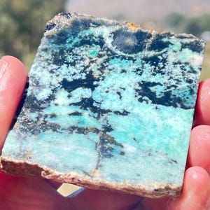 Può includere: Una lastra rettangolare di pietra naturale con una straordinaria tavolozza di colori. La pietra presenta un mix di tonalità turchesi, nere e marroni, creando un aspetto unico e strutturato. La pietra è tenuta in mano, mostrando la sua bellezza naturale.