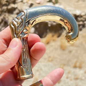 May include: A gold-toned metal cane handle with an ornate design. The handle is shaped like a hook and has a detailed, textured surface.