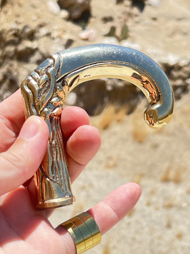 May include: A gold-toned metal cane handle with an ornate design. The handle is shaped like a hook and has a detailed, textured surface.