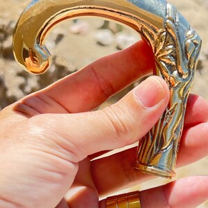 May include: A gold-toned metal cane handle with an ornate design. The handle is shaped like a hook and has a textured surface.