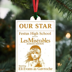 May include: A rectangular Christmas ornament with the text "OUR STAR" and "Les Misérables SCHOOL EDITION." The ornament features an illustration of a child and the text "Starring Eli Evans as Gavroche March 20-23, 2025."