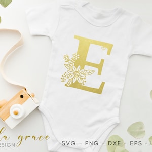 May include: A white onesie with a gold letter E and floral design. The letter E is surrounded by small flowers and leaves.