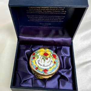 May include: A decorative, round enamel box with a lid featuring a floral design and the words "Happy Anniversary." The box is in a dark blue presentation box with a satin lining. The box is from Alastor Enamels, England.